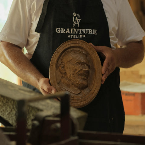 Dieses Bild zeigt eine Person, die eine blaue Jeansschürze mit dem Logo "Grainture Atelier" trägt und eine detaillierte Holztafel mit dem geschnitzten Reliefporträt eines bärtigen Mannes hält. Die Person steht in einer Werkstatt, mit unscharfen Holzbearbeitungswerkzeugen und Materialien im Vorder- und Hintergrund. Die natürliche Beleuchtung hebt die komplizierten Details und die Textur der Schnitzerei hervor und unterstreicht die handwerkliche Qualität der Arbeit.