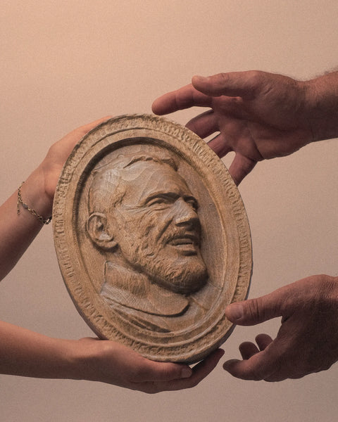 This image shows a carved wooden plaque being passed between two people. The plaque features a detailed relief portrait of a bearded man. The background is plain, focusing attention on the plaque and the hands exchanging it. The lighting is soft, highlighting the texture of the wood and the intricate details of the carving. The left hand appears to belong to a younger person, while the right hand seems to belong to an older individual, suggesting a transfer of heritage or legacy.