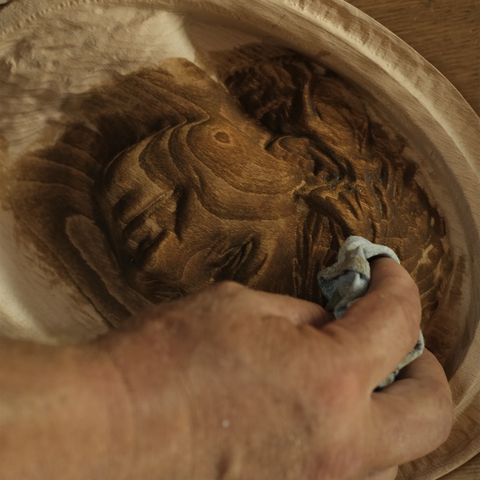 Dieses Bild zeigt eine Nahaufnahme einer Hand, die eine Beize oder ein Finish auf eine Holztafel mit einem geschnitzten Reliefporträt aufträgt. Der Hintergrund ist leicht unscharf, was auf eine Werkstattumgebung schließen lässt. Die warme Beleuchtung hebt die komplizierten Details und die Tiefe der Schnitzerei hervor und unterstreicht die sorgfältige handwerkliche Arbeit und den letzten Schliff bei der Herstellung des Kunstwerks.