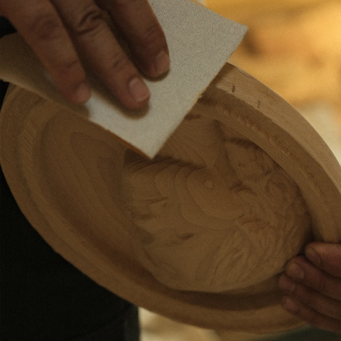 Dieses Bild zeigt eine Nahaufnahme von zwei Händen, die an einem hölzernen Flachrelief mit einem geschnitzten Reliefporträt arbeiten. Eine Hand hält die Platte fest, während die andere mit einem Stück Sandpapier die Oberfläche glättet, um den letzten Schliff bei der Holzschnitzerei zu betonen. Der Hintergrund ist leicht verschwommen und suggeriert eine Werkstattumgebung mit warmer Beleuchtung, die die komplizierten Details und die Textur der Schnitzerei hervorhebt. 