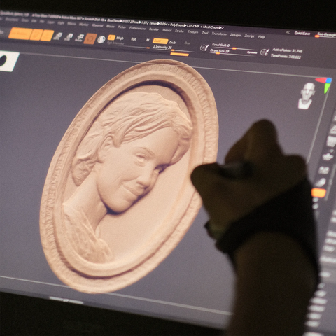Dieses Bild zeigt ein digitales Rendering eines geschnitzten Reliefporträts einer Frau auf einem Computerbildschirm. Eine Hand, die einen Handschuh trägt und einen Stift hält, ist bei der Arbeit auf dem Tablet zu sehen, was die digitale Kunstfertigkeit und Präzision bei der Erstellung des Entwurfs unterstreicht. Die Beleuchtung ist gedämpft und unterstreicht die modernen Techniken der Holzschnitzerei.